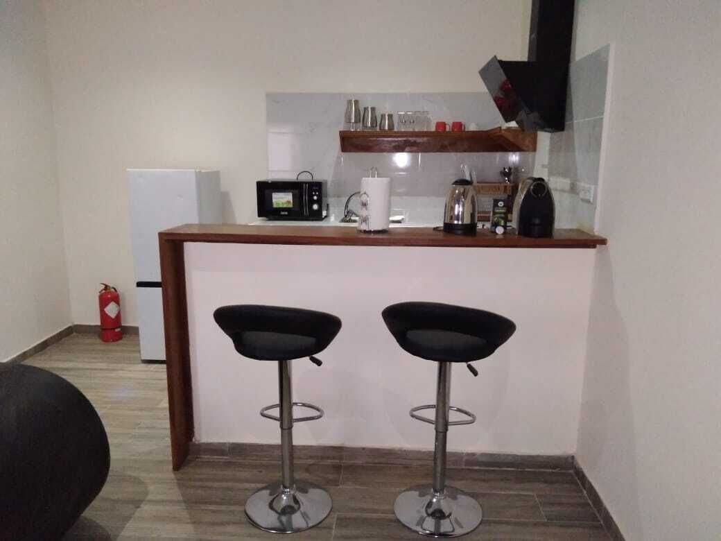 A compact kitchen area with a microwave, toaster, and coffee maker on the counter. There are two black bar stools in front of the counter. A fire extinguisher is visible in the background.