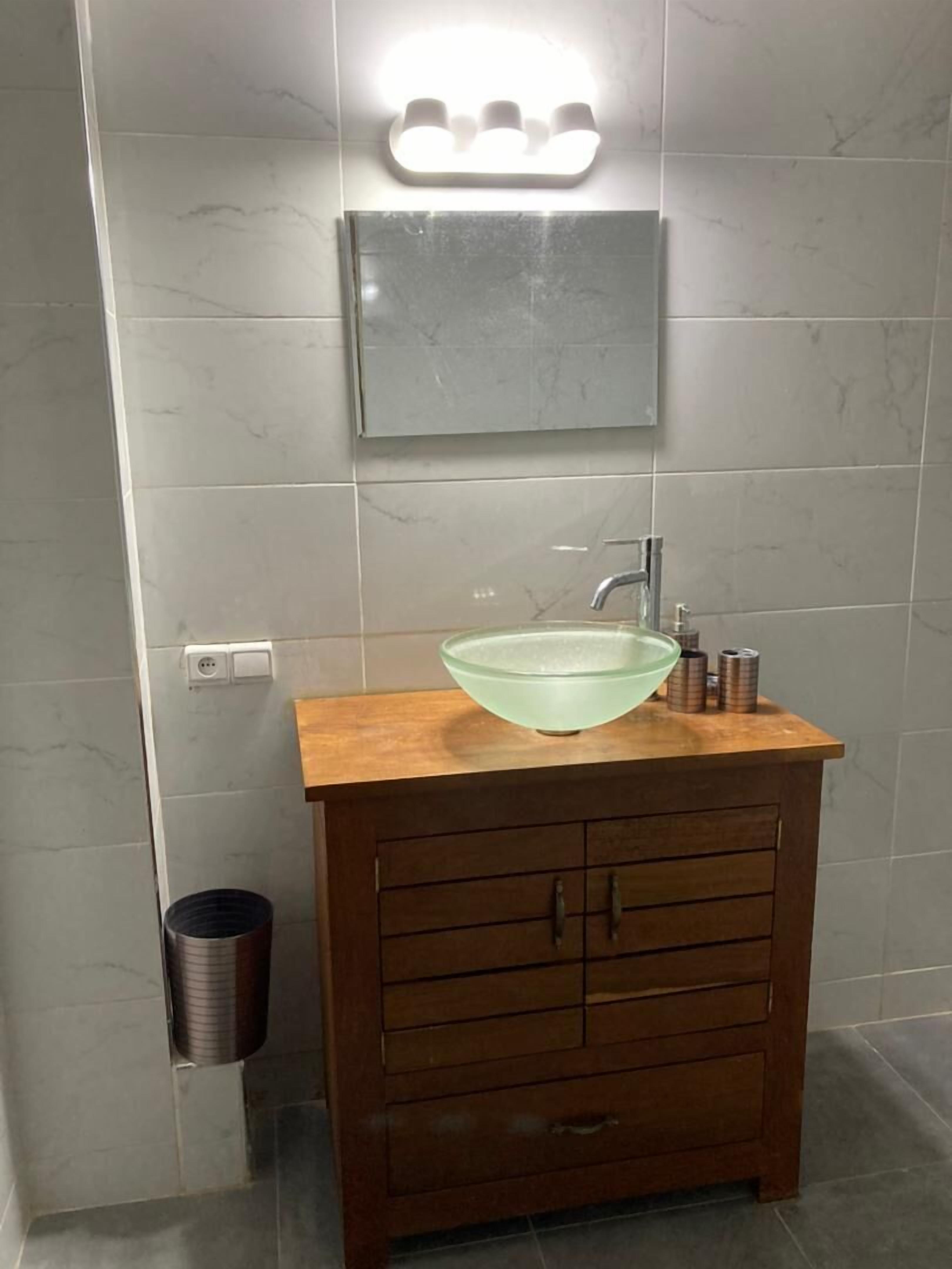 A bathroom with a wooden vanity, a glass bowl sink, a mirror, and a trash can.