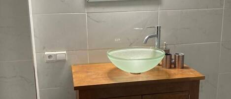 A bathroom with a wooden vanity, a glass bowl sink, a mirror, and a trash can.