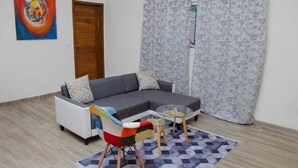 A living room with a grey sectional sofa, a colorful chair, a glass coffee table, and patterned curtains. - Cap Skirring (Cap Skirring)