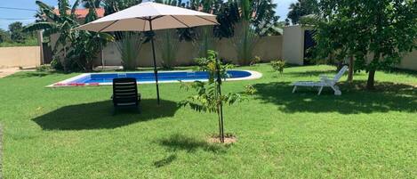 A backyard with a swimming pool, a covered seating area, and a small tree.