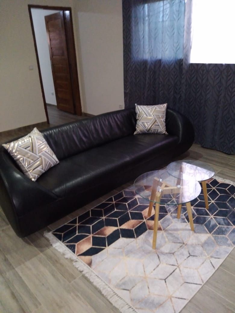 A living room with a black leather sofa, two patterned cushions, a glass coffee table with wooden legs, and a geometric patterned rug.