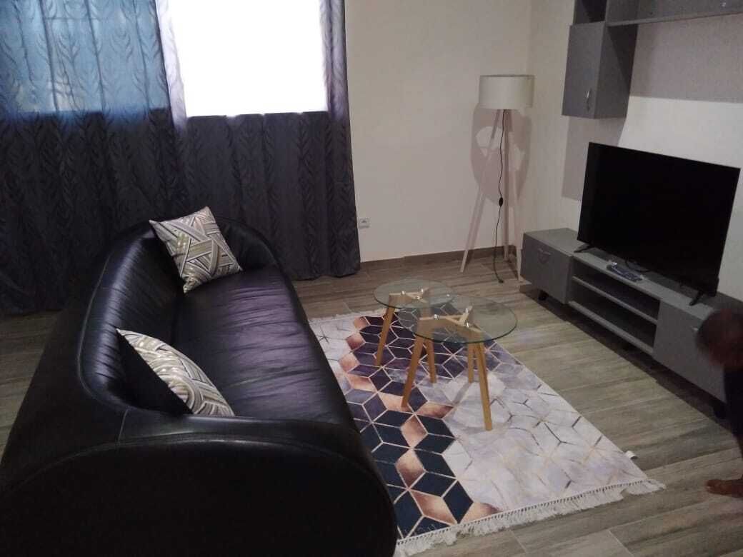 A living room with a black leather sofa, a glass coffee table, a patterned rug, a standing lamp, and a flat-screen TV.
