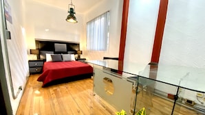 A modern bedroom with a glass-enclosed balcony, a wooden floor, a red bedspread, and a large potted plant.