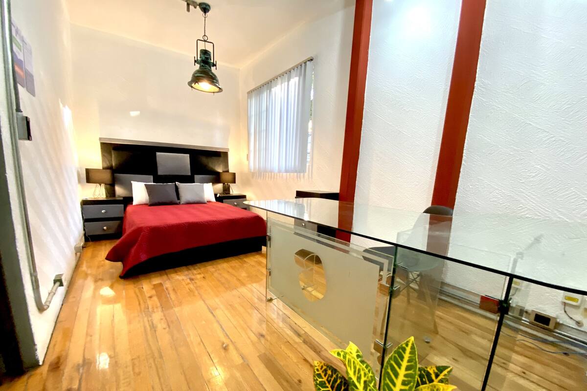 Ein modernes Schlafzimmer mit einem verglasten Balkon, Holzboden, roter Bettdecke und einer großen Topfpflanze.