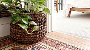Huoneessa on sänky, punottu kori, jossa on kasveja, ja kuviollinen matto.