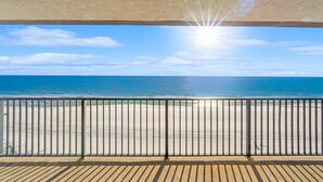 Une vue sur la plage depuis un balcon doté d’une rambarde en métal, avec un ciel dégagé et un soleil éclatant.