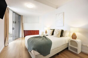 A modern bedroom with a large bed, a sliding glass door, a wooden headboard, a bedside table with a lamp, and a wall-mounted artwork.
