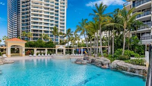 A resort pool with clear blue water, surrounded by palm trees and a large hotel building in the background. - Surfers Paradise: Family Fun w/ 3 Pools, Gym & Spa (Surfers Paradise)