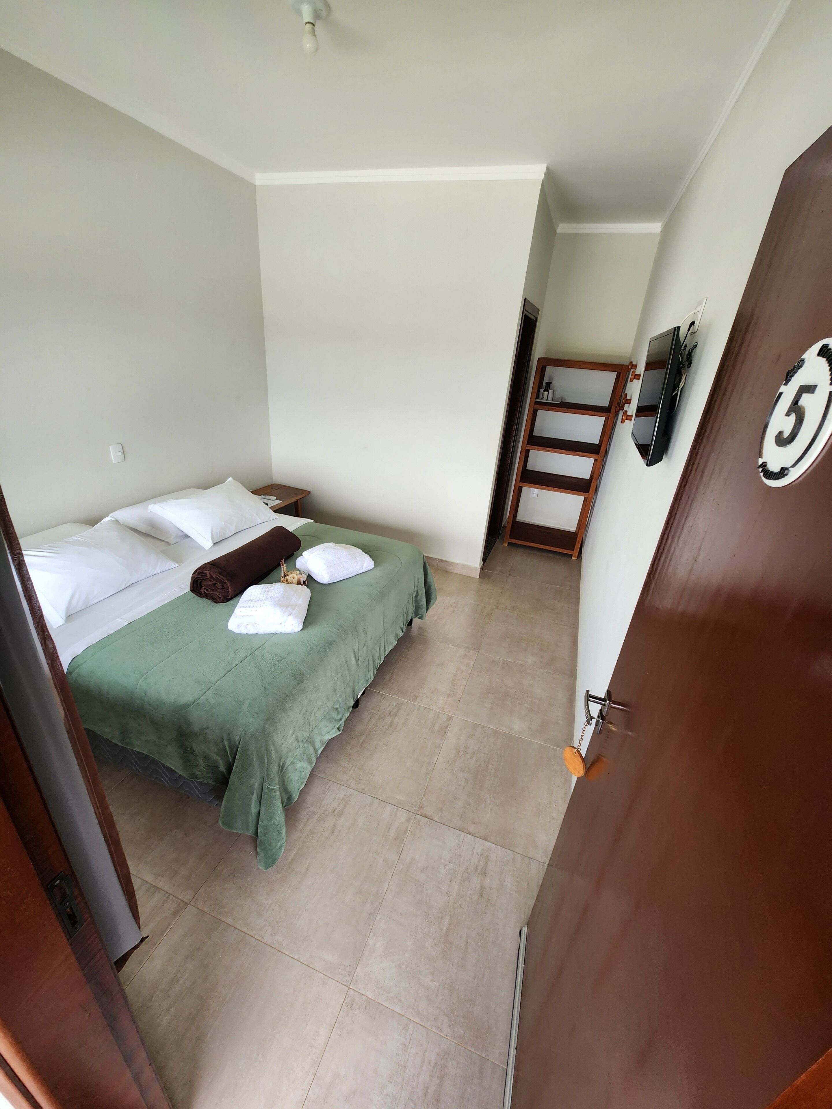 Une chambre d’hôtel avec un lit, une couvre-lit verte, des oreillers blancs et une couverture marron. On y trouve une porte en bois avec une plaque numérotée, un téléviseur fixé au mur et une échelle en bois appuyée contre le mur.