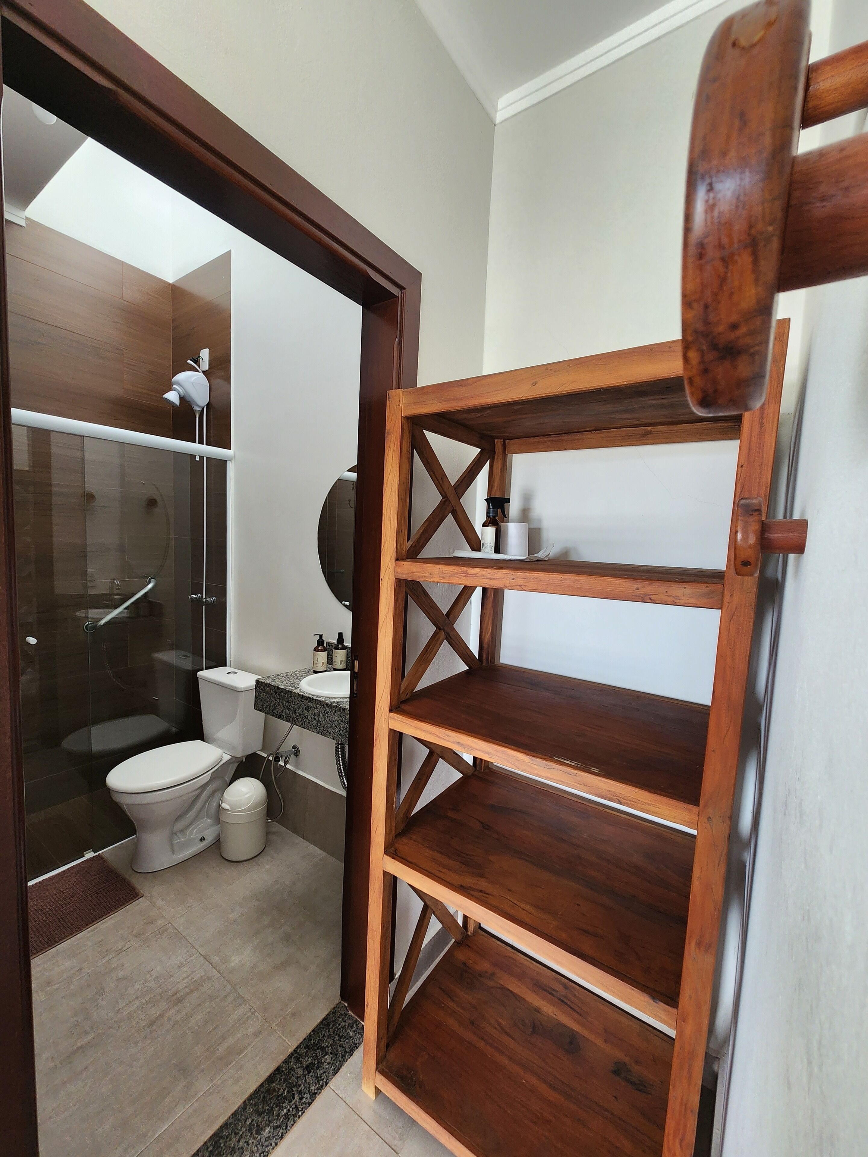 Baño con cabina de ducha de vidrio, inodoro blanco, estante de madera y lavamanos blanco sobre encimera de granito.