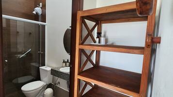 Baño con cabina de ducha de vidrio, inodoro blanco, estante de madera y lavamanos blanco sobre encimera de granito.