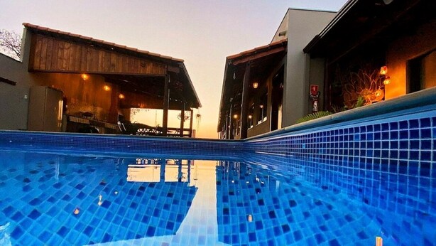 A swimming pool with blue tiles, surrounded by buildings with wooden structures and a covered area. - Pousada Luz de Candeia (São João Batista do Glória)