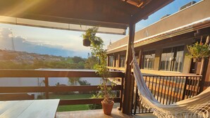 A wooden deck with a hammock and potted plants, overlooking a residential area with houses and a body of water. - Pousada Luz de Candeia (São João Batista do Glória)
