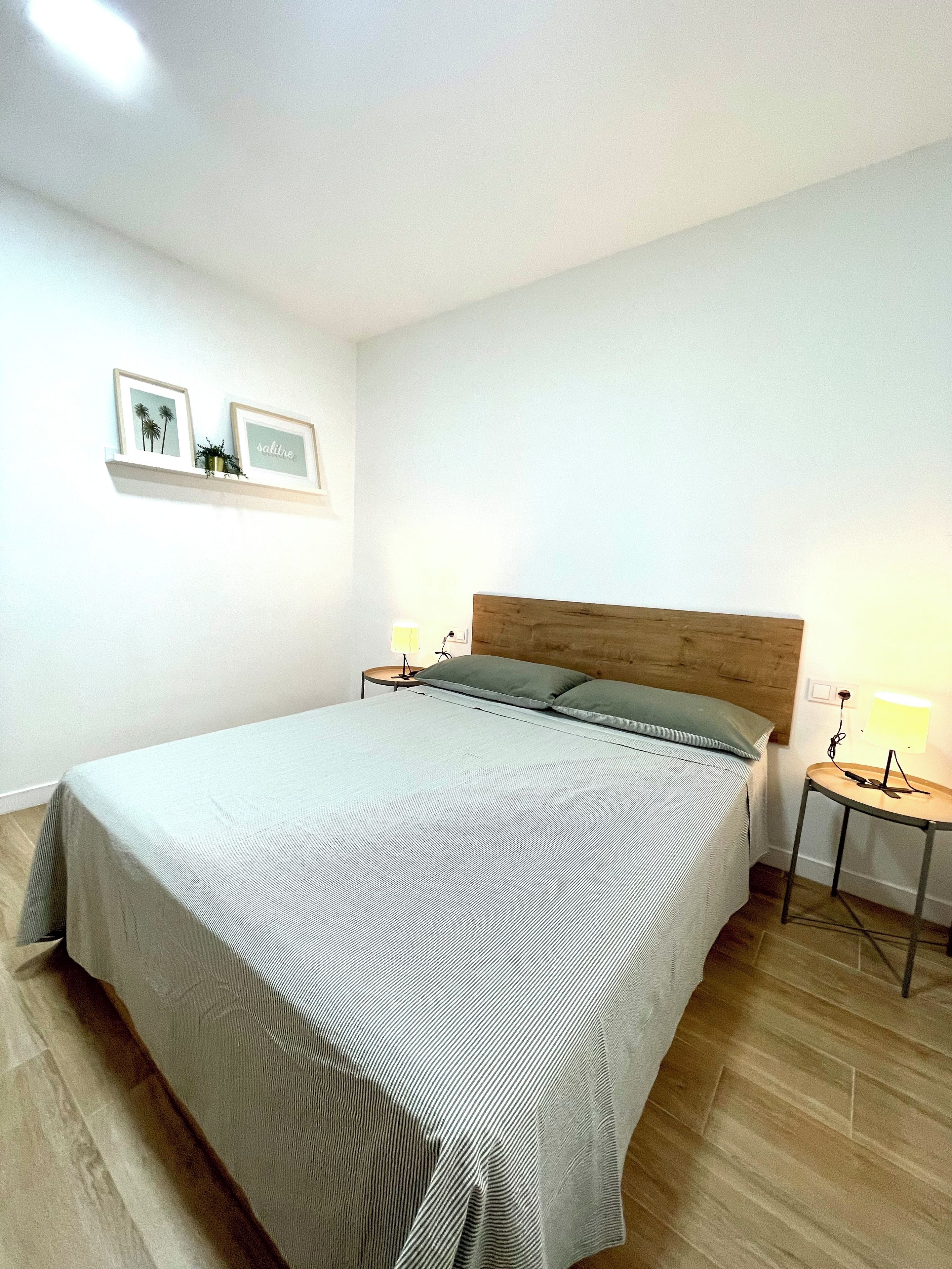 A single bed with a wooden headboard, a bedside table with a lamp, and a wall-mounted shelf with two framed pictures.