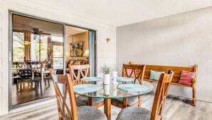 Een eetgedeelte met een glazen tafel en houten stoelen, een bank met kussens, en uitzicht op de aangrenzende woonkamer door een glazen deur.