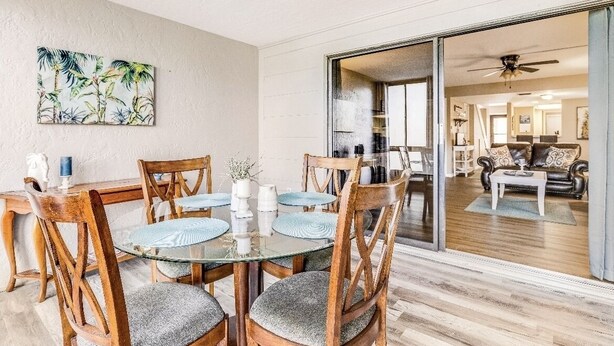 Een eetgedeelte met een glazen tafel en houten stoelen, een keuken zichtbaar door een glazen deur, en een woonkamer met een bank en salontafel.