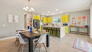 A modern kitchen with yellow cabinets, a central island, and a dining area with a dark wooden table and white chairs. - Kaleidoscope by Avantstay Colorful Escape + Pool (Indio)