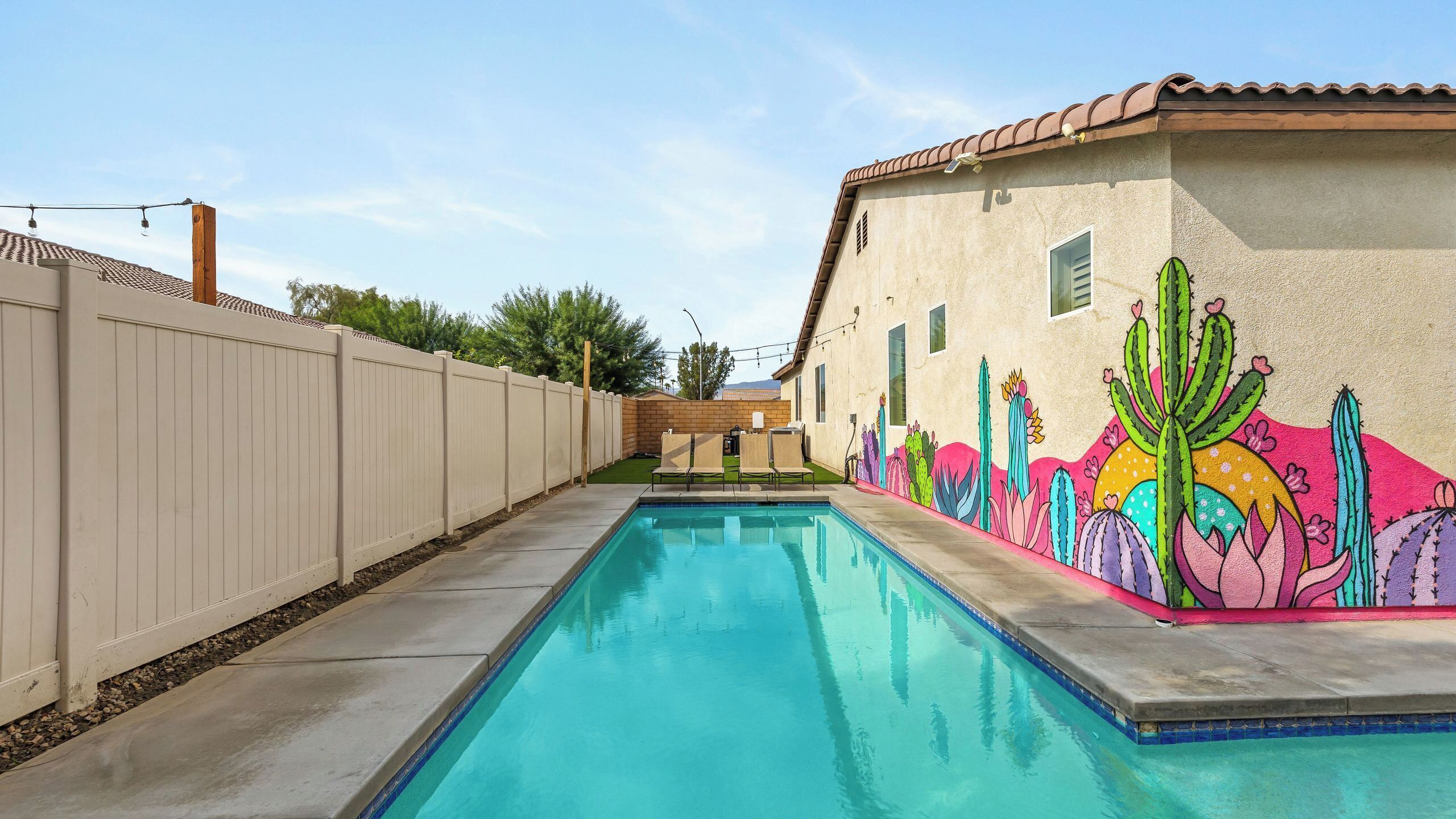 A swimming pool with a cactus mural on one side, a wooden fence, and a clear sky.
