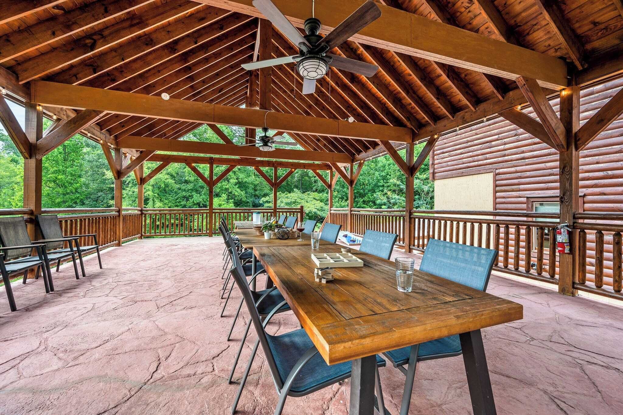 A covered outdoor area with a wooden table and chairs, a ceiling fan, and a view of a forested area.