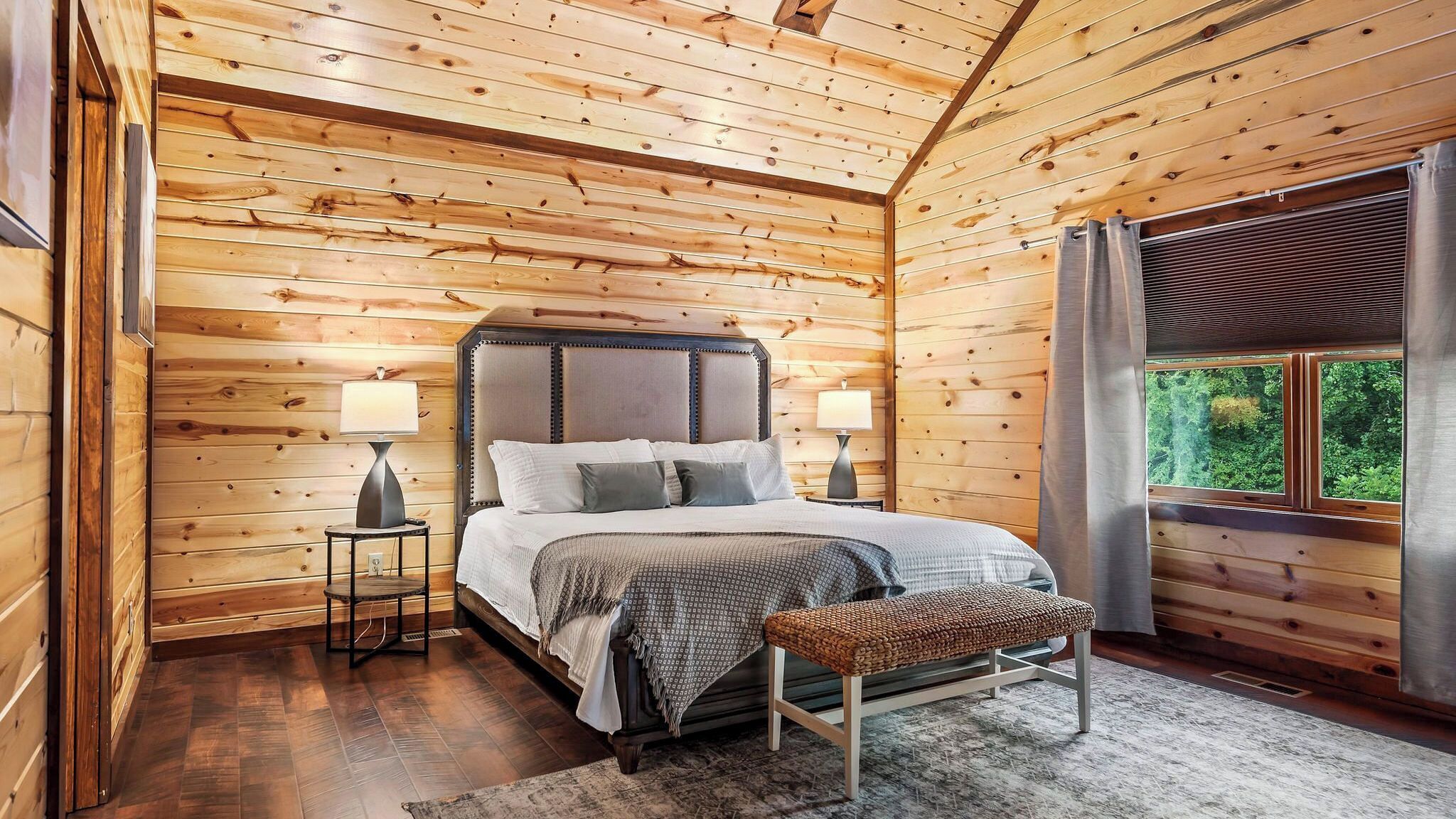 Une chambre dans une cabane en bois, avec un lit, deux tables de chevet avec des lampes, un banc et une fenêtre avec des rideaux.