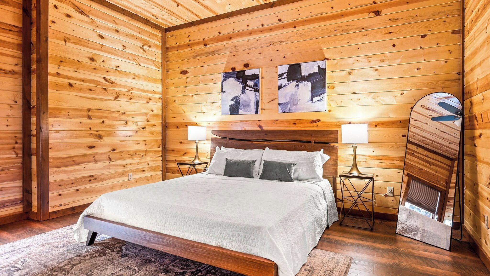 A wooden-paneled room with a bed, two wall-mounted paintings, a ceiling fan, and a nightstand with a lamp.