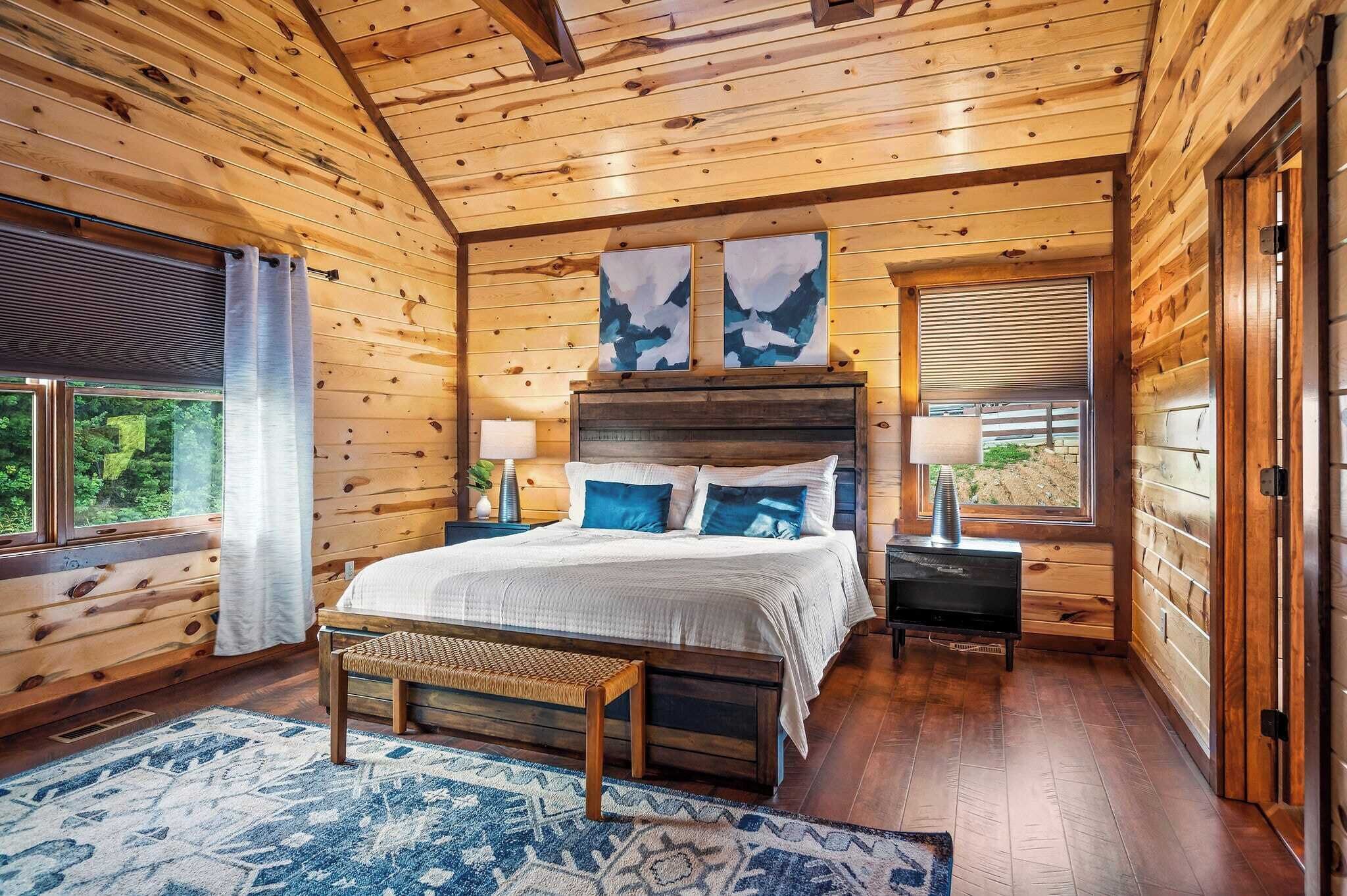 A wooden cabin bedroom with a bed, bench, artwork, and a window with blinds.
