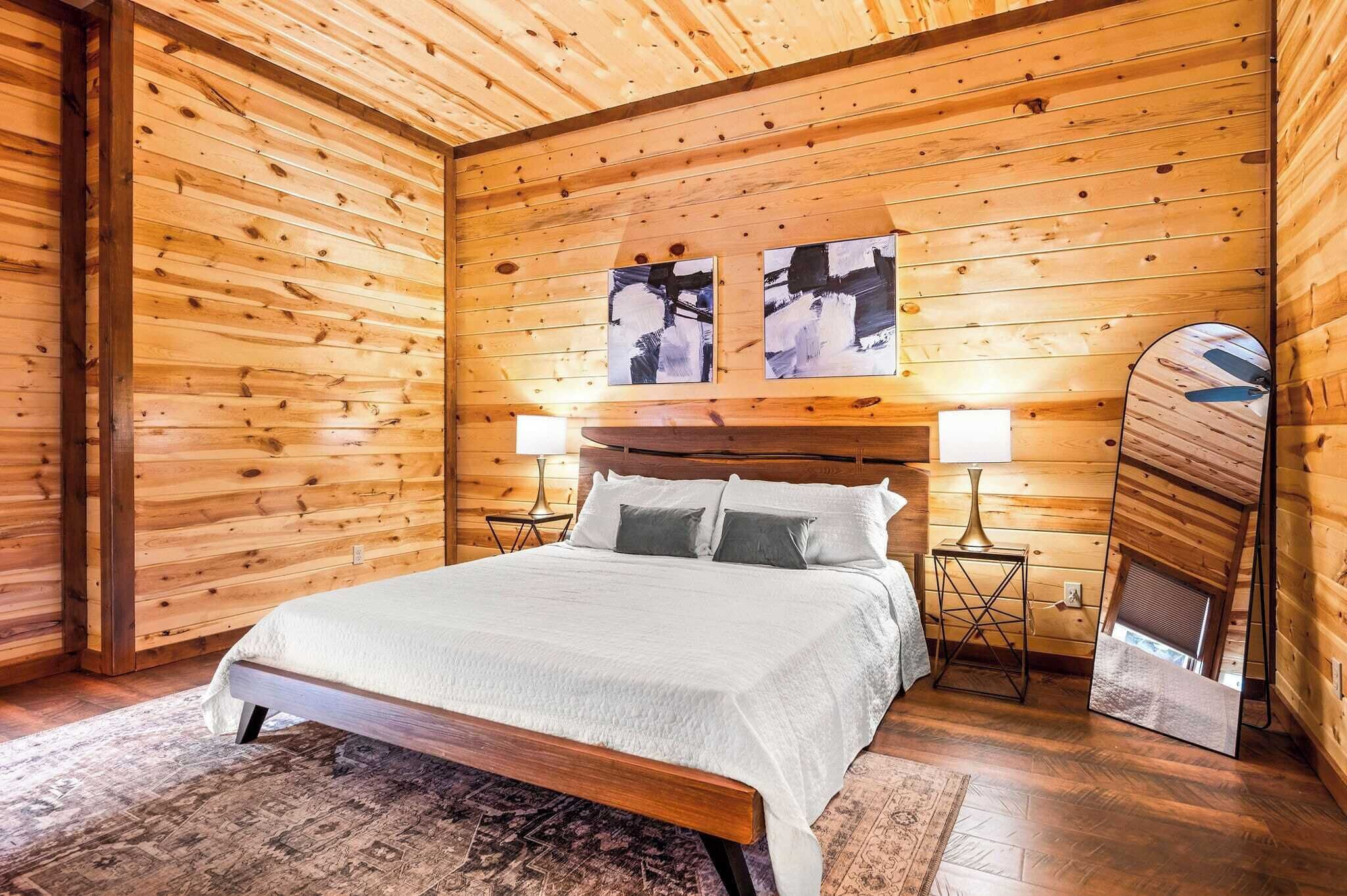 A wooden-paneled bedroom with a bed, two wall-mounted lamps, and two abstract paintings.