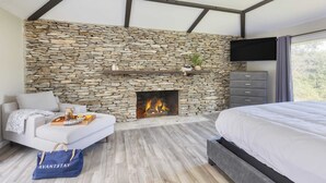 A modern bedroom with a stone fireplace, a sofa, a bed, and a TV mounted on the wall. - Amber Refresh by Avantstay Pool and Hot Tub (New Market)