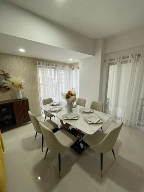 A dining area with a white table set for six, surrounded by six chairs. There is a vase with flowers on the table, and a cabinet with decorative items and glasses on the left.