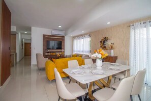 A modern living room with a yellow sofa, a dining table set for four, and a flat-screen TV mounted on the wall.