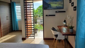 A bedroom with a bed, a desk with a lamp, a TV on the wall, and a view of a garden through an open door.