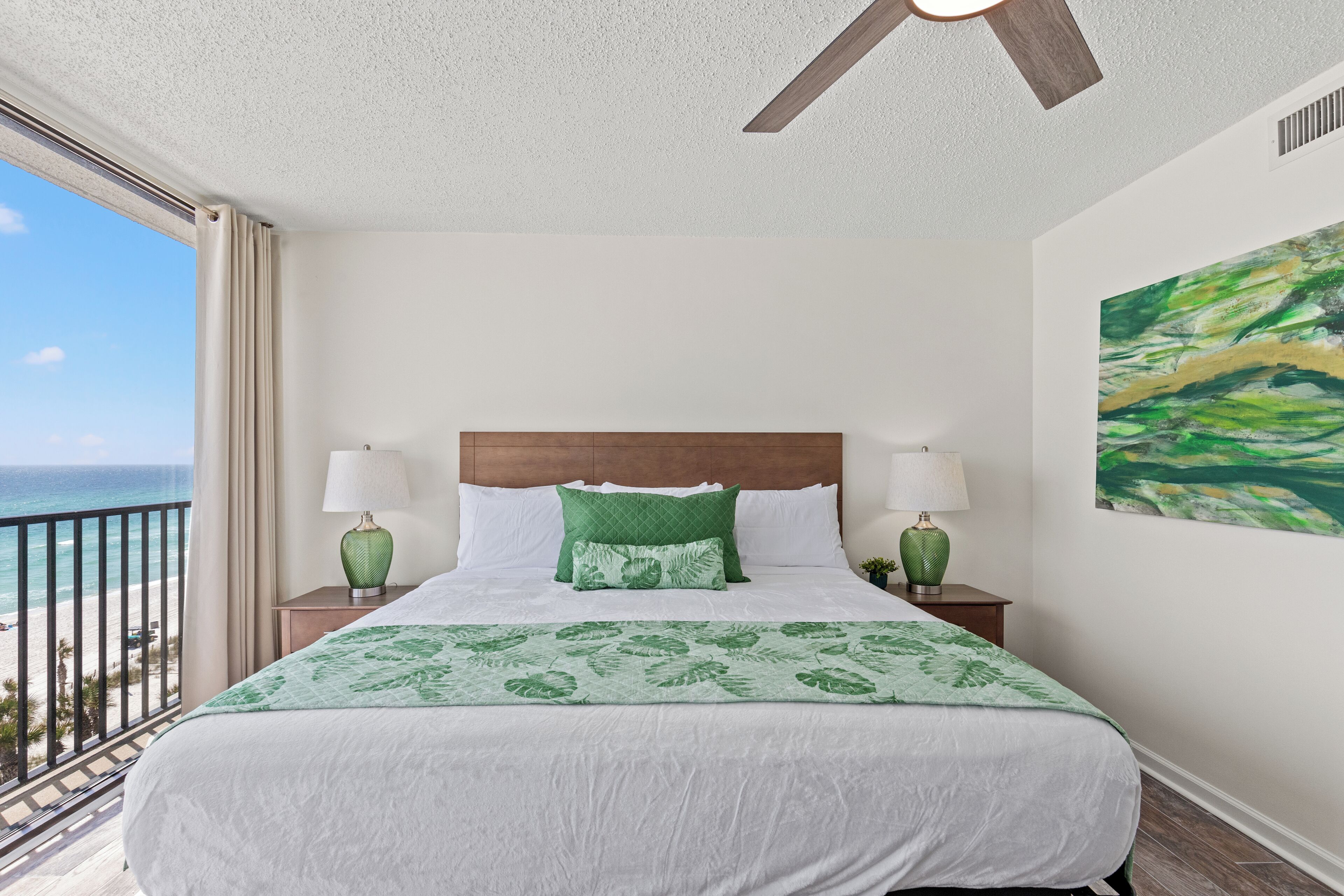 A hotel room with a large bed, two bedside tables with lamps, a view of the beach, and a ceiling fan.
