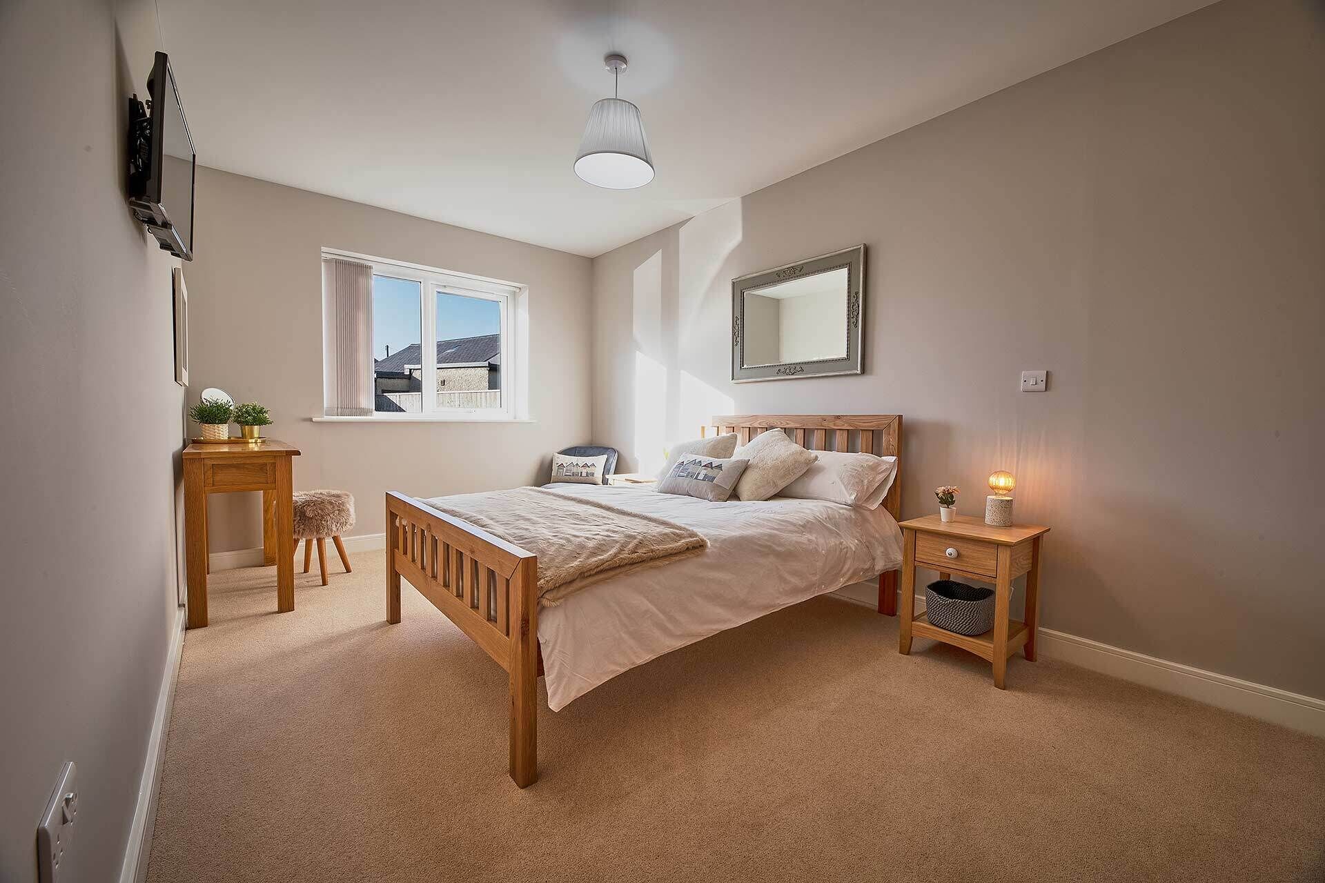 A bedroom with a wooden bed, a bedside table, a nightstand, a mirror, and a window with a view of buildings.