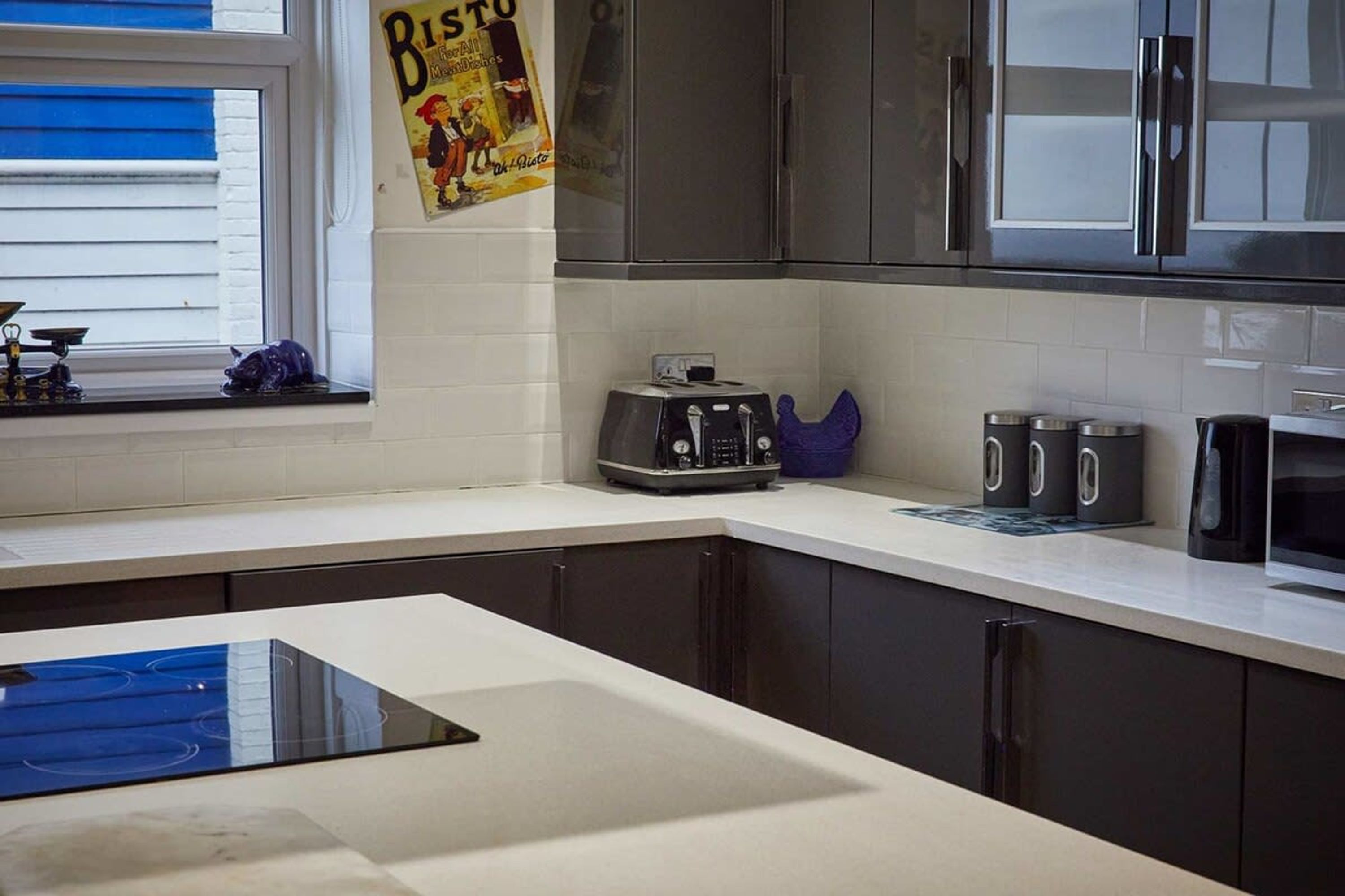 Eine moderne Küche mit dunklen Schränken, einer weißen Arbeitsplatte, einem Toaster, einer Kaffeemaschine und einem Fenster mit Jalousien.