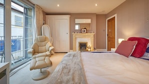 Une chambre avec un grand lit, un fauteuil, une cheminée et une vue sur l’extérieur.