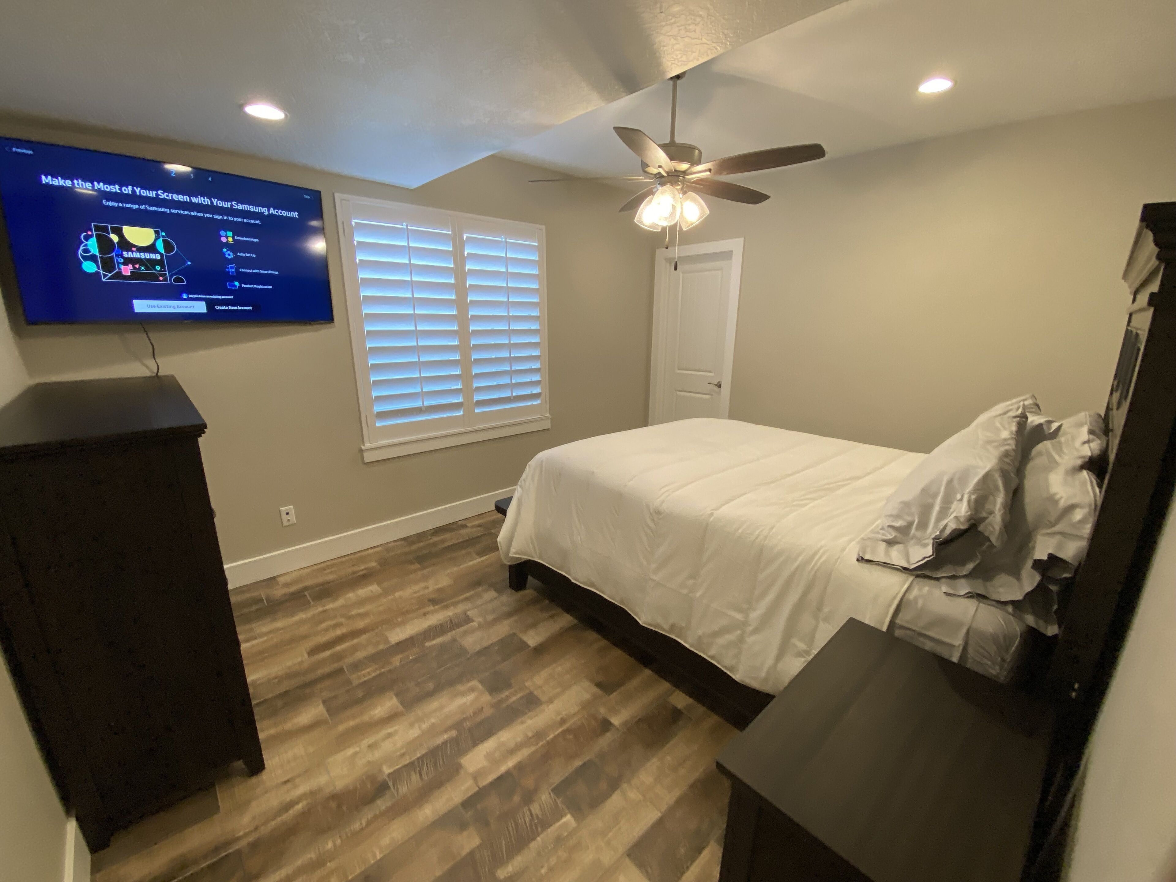 Un dormitorio con cama, televisor montado en la pared, ventilador de techo y ventana con persianas.