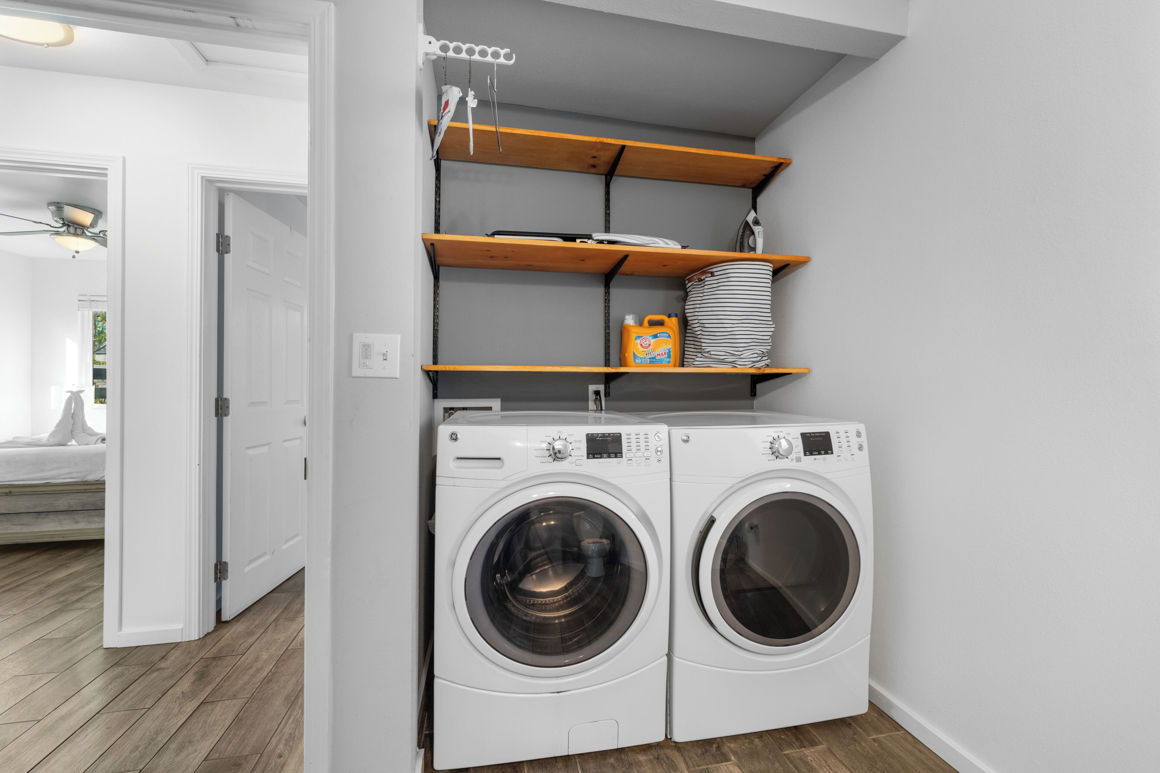 A laundry area with two white front-loading washing machines, wooden shelves above, and a white door leading to another room.