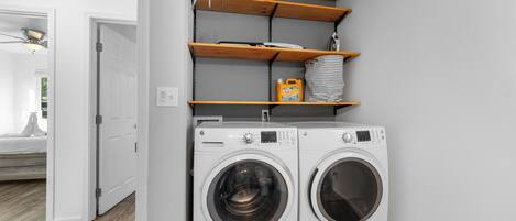 A laundry area with two white front-loading washing machines, wooden shelves above, and a white door leading to another room.