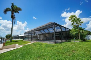A modern glass-enclosed structure with a flat roof, surrounded by well-maintained grass and a few trees. - VRCC Villa Cape Escape (Cape Coral)