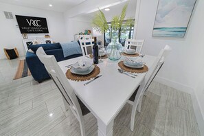 A modern dining area with a white table set for four, surrounded by white chairs. A large mirror reflects the room, and a TV displaying 'VACATION RENTALS CAPE CORAL' is mounted on the wall. - VRCC Villa Cape Escape (Cape Coral)