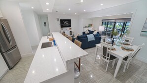Una cocina moderna con una isla central, un comedor con mobiliario blanco y una sala de estar con un sofá azul y un televisor mostrando 'Vacation Rentals Cape Coral'.