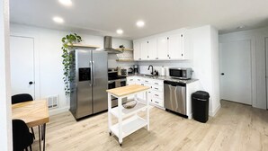 Uma cozinha moderna com armários brancos, eletrodomésticos de aço inoxidável, uma mesa de madeira e uma lixeira preta.