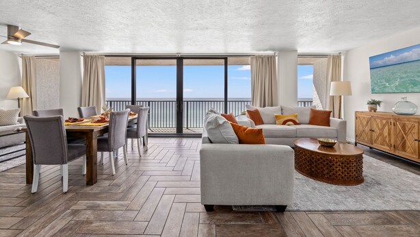 Ampla sala de estar com mesa de jantar, sofás cinzas e vista para o oceano através de grandes janelas.