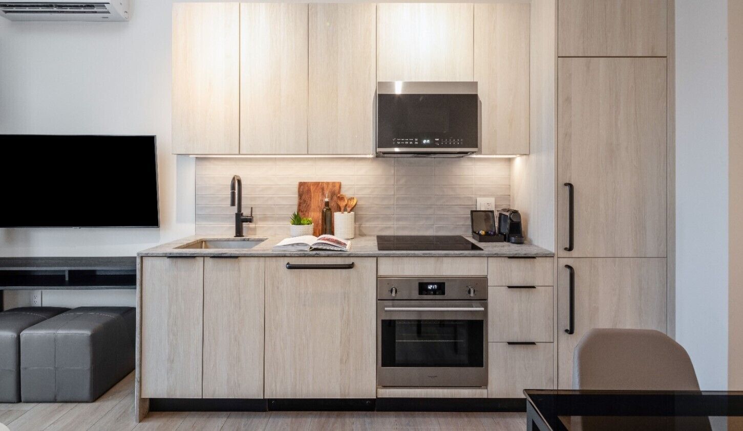 A modern kitchen with light wood cabinetry, a stainless steel range hood, a built-in oven, and a flat-screen TV mounted above the counter.