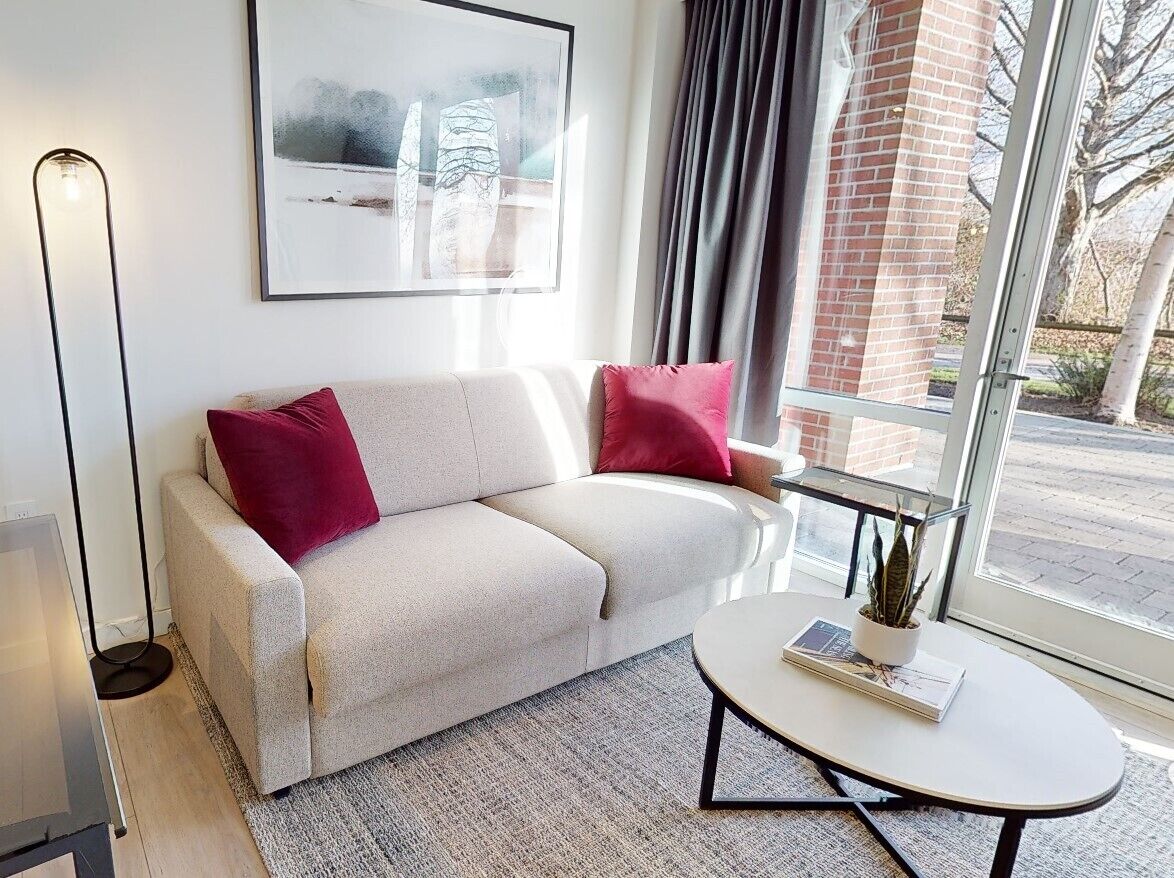 Un salon moderne avec un canapé beige, deux coussins rouges, une table basse ronde sur laquelle se trouvent un livre et une plante, un lampadaire et une grande fenêtre donnant sur un bâtiment en briques et des arbres.