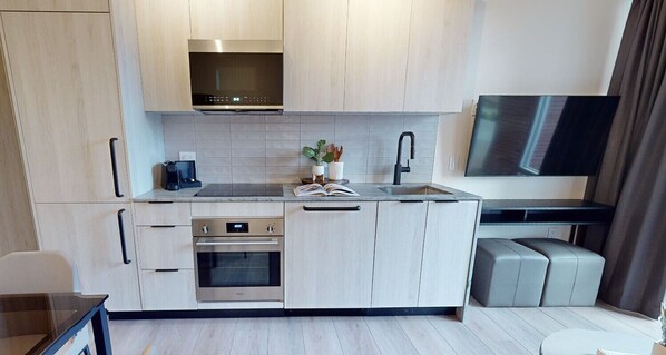 A modern kitchen with a built-in oven, a microwave, and a stainless steel range hood. There is a small dining area with a table and chairs, and a flat-screen TV mounted on the wall.