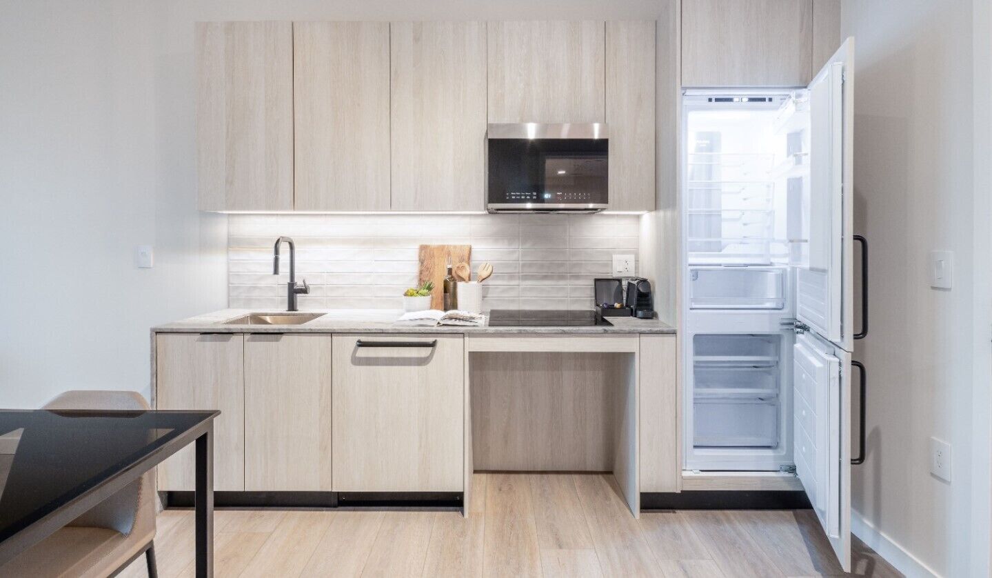 A modern kitchen with light wood cabinets, a stainless steel microwave, a built-in oven, and a white refrigerator.