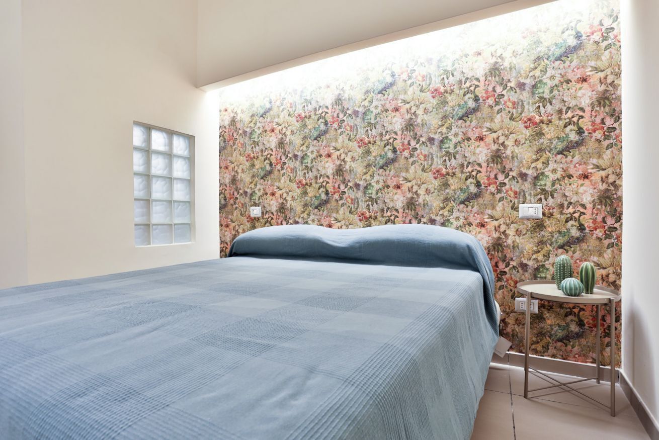 Een slaapkamer met bloemenbehang, een bed met een blauwe spreidek, een tafeltje met twee cactussen en een raam met geëtste glas.