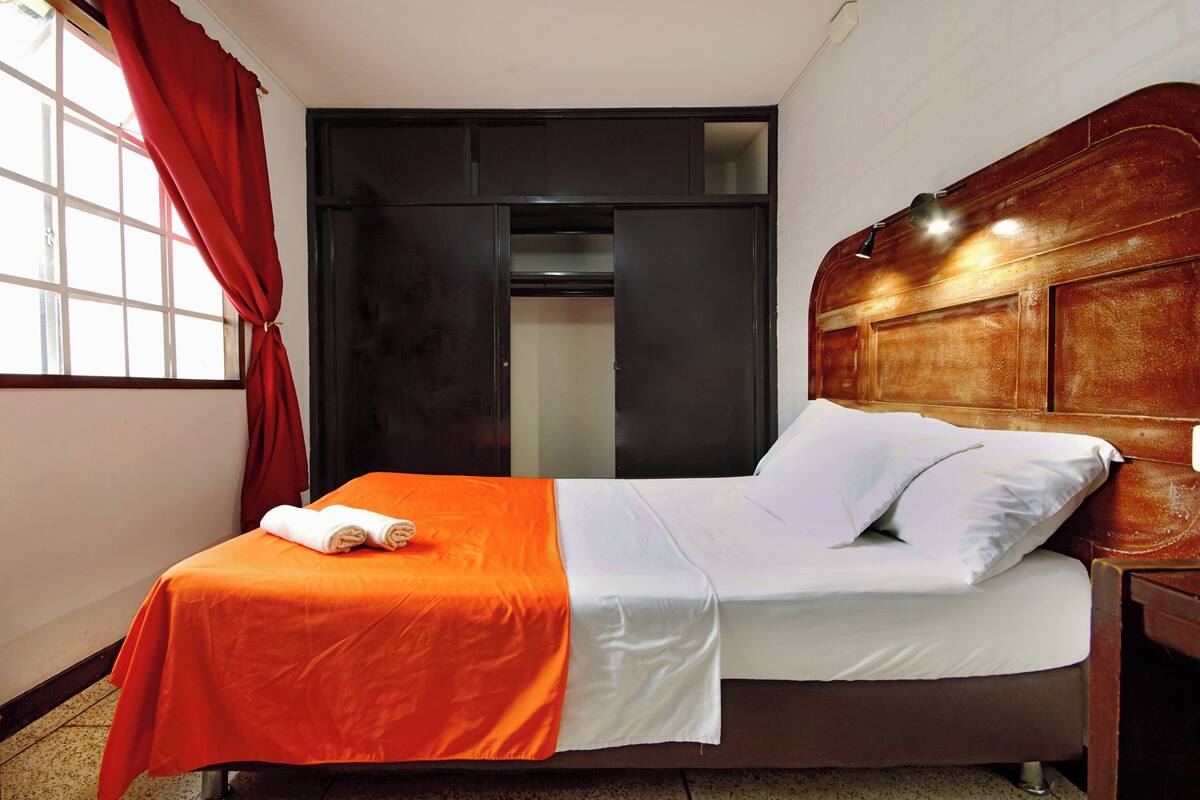Une chambre d’hôtel avec un lit, une tête de lit en bois, une fenêtre avec des rideaux rouges et une porte donnant sur un placard.